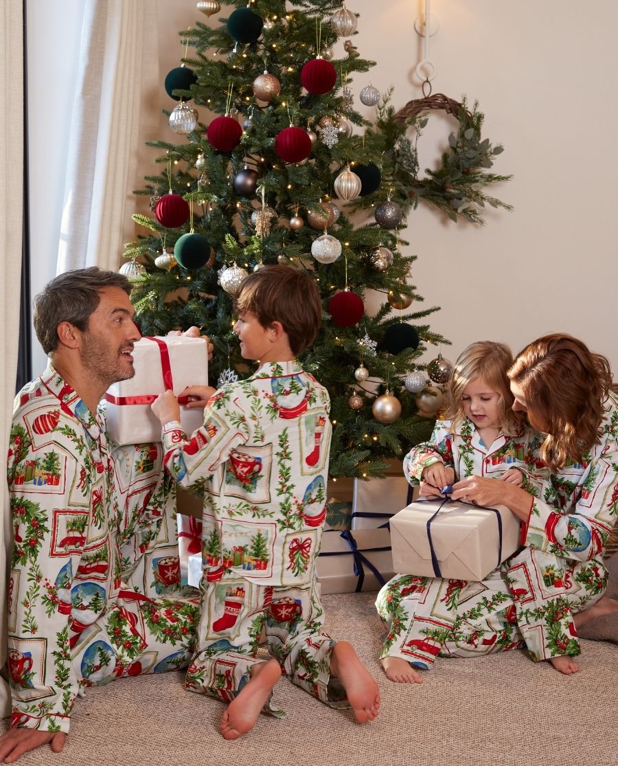 A family of four wearing matching Christmas-themed pajamas sits on the floor in front of a decorated Christmas tree, opening wrapped presents in a cozy living room setting.