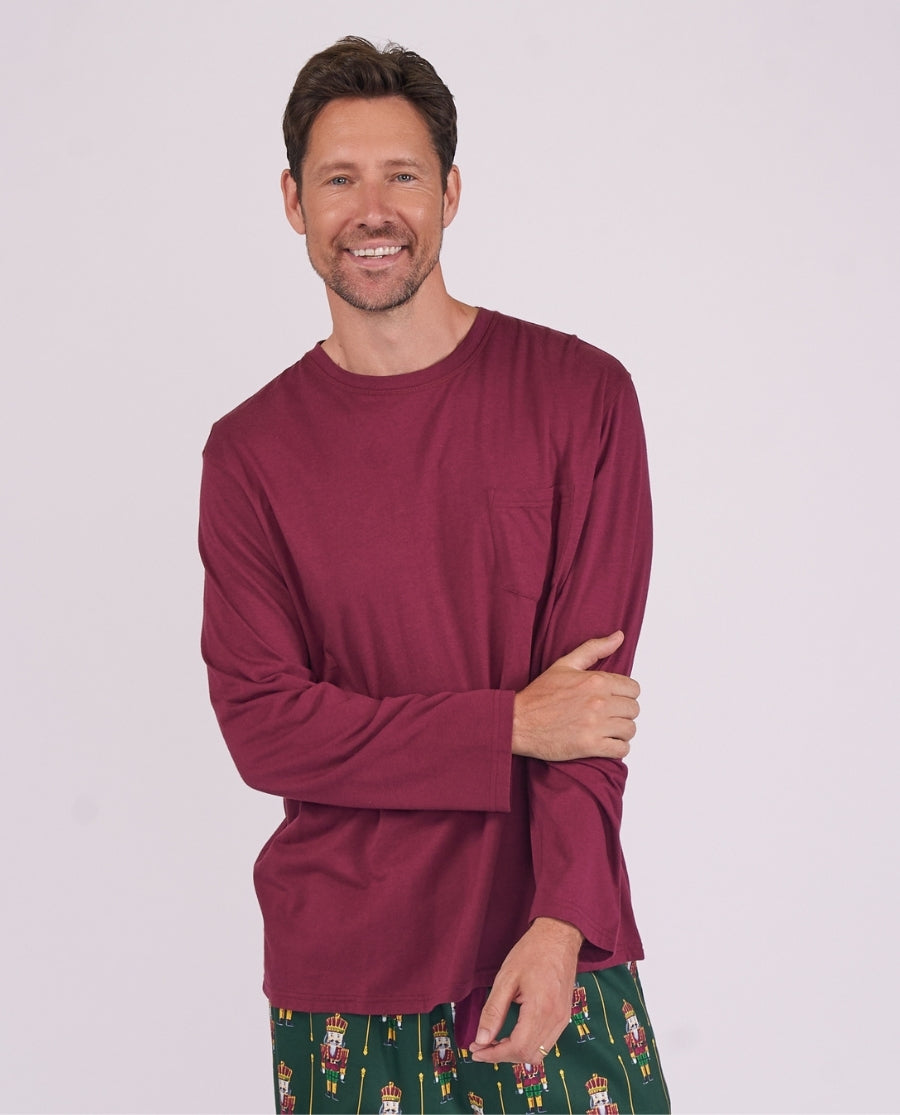 A smiling man wearing a long-sleeve crewneck shirt in burgundy with a chest pocket, paired with green patterned pajama pants, against a light gray studio background.