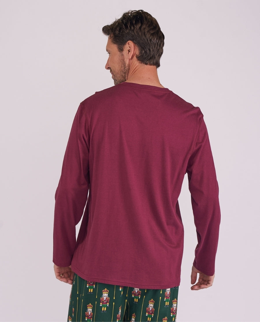 A man shown from the back wearing a long-sleeve burgundy shirt and green patterned pajama pants with nutcracker illustrations, against a plain light gray studio background.